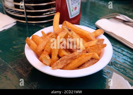 Up close image of amall white bowl with steak fries or French fries on a green picnic table and a blurred ketchup bottle in background; side dish. Stock Photo