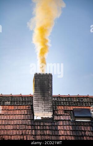 A thick yellow smoke rises to the blue sky from the chimney of the family house. The chimney smokes from the morning ignite in the stove. Stock Photo