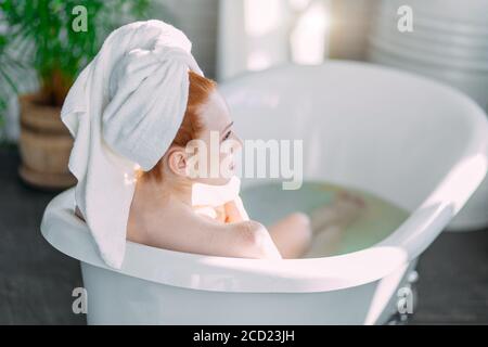 Young woman with towel on head getting spa treatment at beauty salon, relaxing in bathtub with green plants and flowers on background. Bodycare and Re Stock Photo