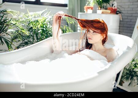Miserable looking disappointed woman taking foam bath holding her wet red hair in hands being extremely upset with hair condition, making grimace diss Stock Photo