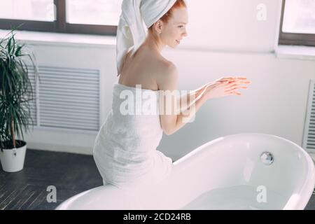 Beautiful woman in bath wear is going to wash her body in the bathtub at home, checking water with hand, enjoying minutes of tranquility and silence Stock Photo