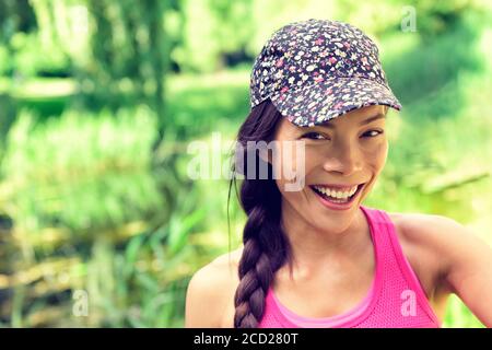 Young happy healthy Asian girl smiling to the camera. Chinese Caucasian multiracial woman wearing fashion cap and hair braid in summer park green Stock Photo