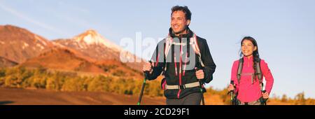 Hiking hikers couple trekking in nature mountains at sunset with walking poles and backpack wearing outdoor gear jackets. Happy multiracial group of Stock Photo