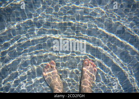 White man's feet in the pool, mixed dark and light gray mosaic floor mixed The waves on the water surface jump in the golden sun. Stock Photo