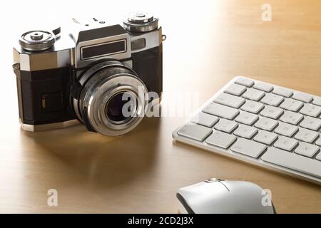 Retro analog SLR camera next to computer keyboard and mouse on wooden desk in office, digital photography or image processing concept, selective focus Stock Photo