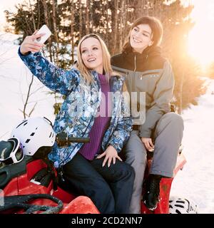 Two cheerful pretty women posing taking selfie against setting sun in snowy mountains sitting on red quad-bike in the winter. Concept of mobile photography and quad biking. Stock Photo