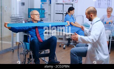Handicapped injured man training with elastic band under strict medical supervision in modern private recovery facility. Invalid physiotherapy program, health care injury rehabilitation Stock Photo