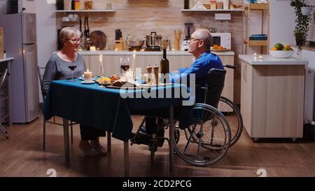 Senior couple coming home for romantic dinner. Old man in wheelchair dining with his cheerful wife sitting at the table in the kitchen. Imobilized paralyzed handicapped husband having romantic dinner Stock Photo
