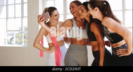 Four fitness women standing at the gym and taking a selfie. Happy fitness women relaxing after workout taking selfie using mobile phone. Stock Photo