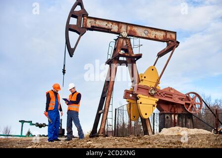 Smiling extraction operator writing on clipboard while working with colleague in oil field near petroleum pump jack. Male workers in helmets, work vests. Concept of oil extraction, petroleum industry. Stock Photo