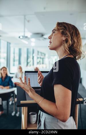 Side view of a businesswoman talking to colleagues from a podium. Female executive addressing to group of professionals at a conference. Stock Photo