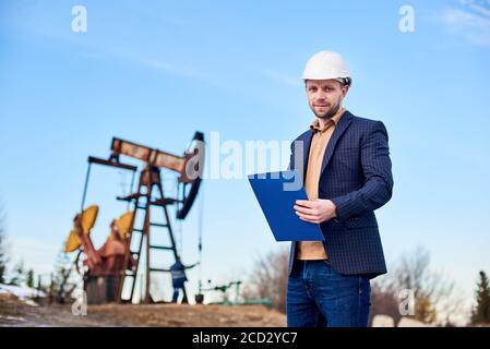 Portrait of engineer wearing suit jacket and white helmet, holding folder and making some notes, oil worker working with oil pump jack and blue sky on background. Concept of petroleum industry Stock Photo