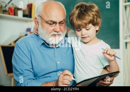 Grandfather with grandson learning together. Friendly child boy with old mature teacher in classroom near blackboard desk. Teacher is skilled leader. Stock Photo