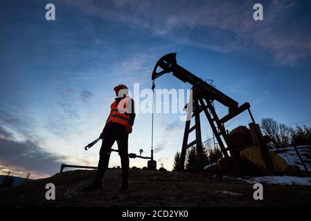 Horizontal snapshot of a silhouette of an oilman in orange vest and helmet, standing with his back to the oil pump jack in oilfield holding a pipe wrench, looking away, evening sky on background Stock Photo