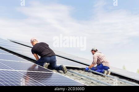 Process of building a solar station. Electrical engineers mans are working installing solar panels on solar station on house roof against blue sky Stock Photo