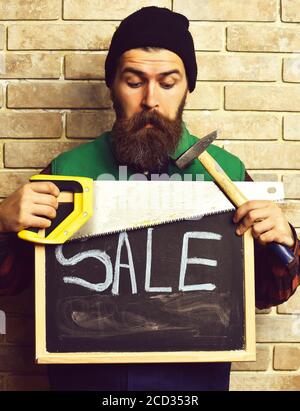 bearded man, long beard, brutal caucasian hipster with moustache holding various building tools: saw, hammer and board with inscription sale, surprised face, brick wall background Stock Photo