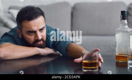 Frustrated man with beard takes a glass of whiskey from table in living room interior Stock Photo