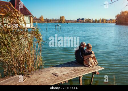 Senior family couple relaxing by autumn lake. Happy man and woman enjoying nature and hugging sitting on pier Stock Photo