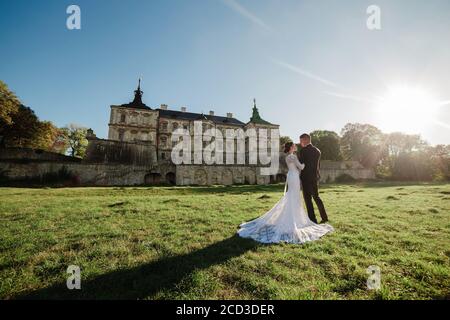 Lovely wedding couple walking in the sunlight near the old castle. Stylish handsome bride and groom in arms against the backdrop of an ancient Stock Photo