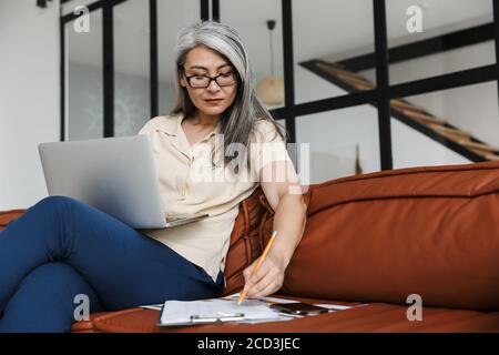 Photo of adult asian woman working with laptop and making down notes while sitting on sofa in living room Stock Photo