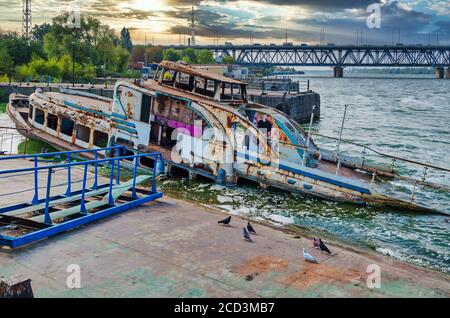 Semi-flooded passenger river ship at abandoned pier on background of sunset and railway bridge Stock Photo