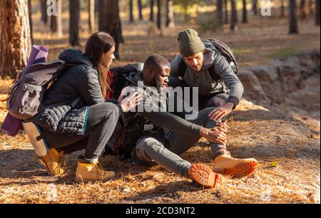 Friends helping black guy with injured leg, hiking at forest Stock Photo