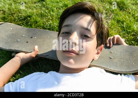 Boy with brown hair lying on grass, head resting on skateboard, looking at camera. Stock Photo