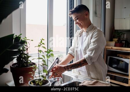 Young woman with short hair standing in kitchen, washing hand in sink. Stock Photo