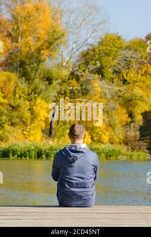 Young man sits on the pier. Autumn sunny day. Back view Stock Photo
