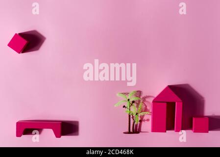 Construction of small house from wooden blocks and cubes, plastic artificial palm on a pink background with shadows, copy space. Top view. Constructor Stock Photo