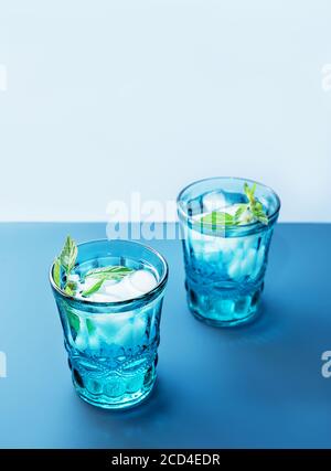 Two beautiful vintage blue glasses with cold drink and ice cubes, with fresh green mint leaves on deep blue and light blue background. Backdrop for your design. Stock Photo