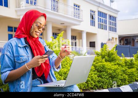 African female sitting outside with her laptop and phone making a payment online Stock Photo