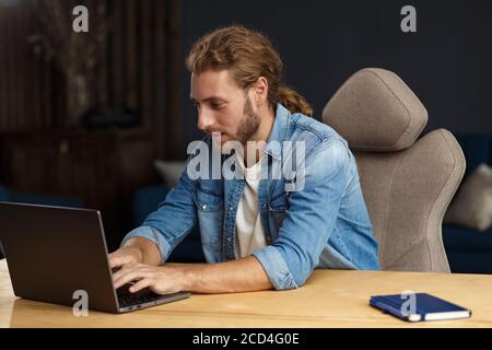 Young handsome curly smiling man with long hair working in home office with laptop. Business portrait of handsome manager sitting at workplace Stock Photo