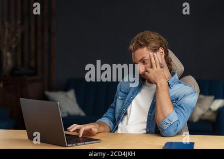 Young handsome curly smiling man with long hair working in home office with laptop. Business portrait of handsome manager sitting at workplace Stock Photo
