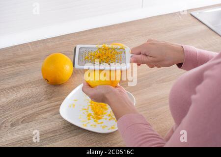 Woman rubbing orange zest with metal grater in the kitchen Stock Photo