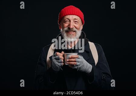 Bearded beggar male smiling, happy even living in street. Cheerful homeless man with mug of hot tea isolated over black background Stock Photo
