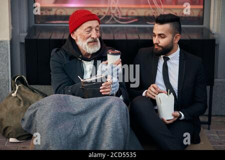 Young beardy man in suit sitting with beggar on floor on street and give cup of coffee. Different segments of society, social inequality Stock Photo