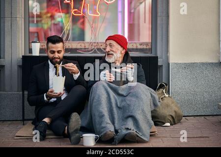 Stylish office worker and beggar male sit eating on street and speaking. Man un tuxedo, poor male in old clothes Stock Photo