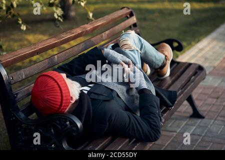 Mature man 70 years old with beard lying on bench at morning, holding cup from coffee for collecting money from citizens Stock Photo