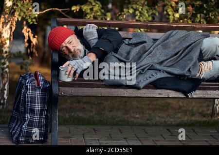 Roofless male lying on street bench asking for money, for any help. Lagguage near bench. Desperate and lonely homeless man without shelter Stock Photo