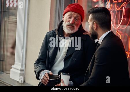 Responsive rich man sat down with homeless man in street and talk, listen to him Stock Photo