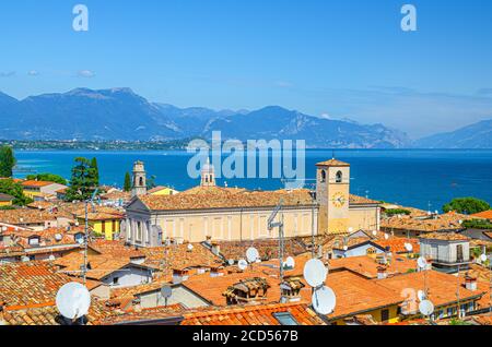 Aerial view of Desenzano del Garda town with bell tower of Duomo di Santa Maria Maddalena Cathedral church, red tiled roof buildings, Garda Lake water surface, mountain range, Lombardy, Northern Italy