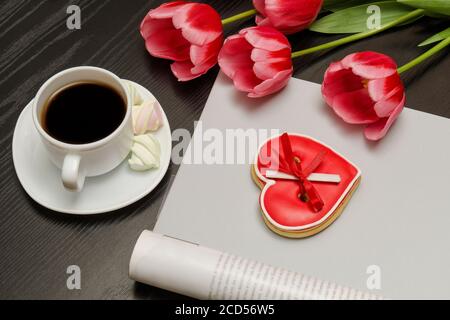 Holiday concept. Bouquet of pink tulips, a cup of coffee, red heart-shaped cookies with a note, empty magazine on a black wooden background. Stock Photo