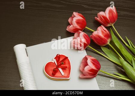 Holiday concept. Bouquet of pink tulips, red heart-shaped cookies with a note, empty magazine on a black wooden background. Directly above Stock Photo