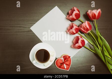 Holiday concept. Bouquet of pink tulips, a cup of coffee, red heart-shaped cookies with a note, a piece of paper on a black wooden background Stock Photo