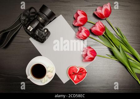 Holiday concept. Bouquet of pink tulips, a cup of coffee, red heart-shaped cookies with a note, a piece of paper on a black wooden background Stock Photo