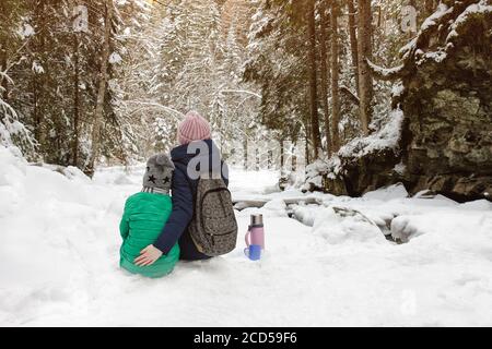 Mom with her son sitting and embracing in the background of snow-covered forest. Winter cloudy day. Back view. Stock Photo