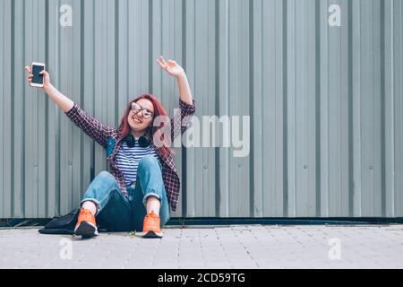 Young beautiful modern smiling female teenager in a checkered shirt and jeans with headphones and smartphone sitting near a wall on the street and Che Stock Photo