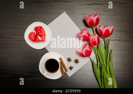 Holiday concept. Bouquet of pink tulips, a cup of coffee, red heart-shaped cookies with a note, a piece of paper on a black wooden background. Directl Stock Photo