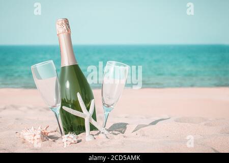 A bottle of champagne and two glasses on a sandy beach with seashells against a blue sea background Stock Photo
